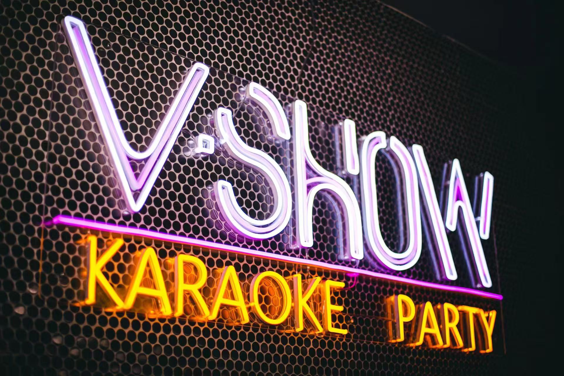 The V Show Karaoke neon sign on a perforated metal wall, reading V Show in white and purple tubing above Karaoke Party in yellow