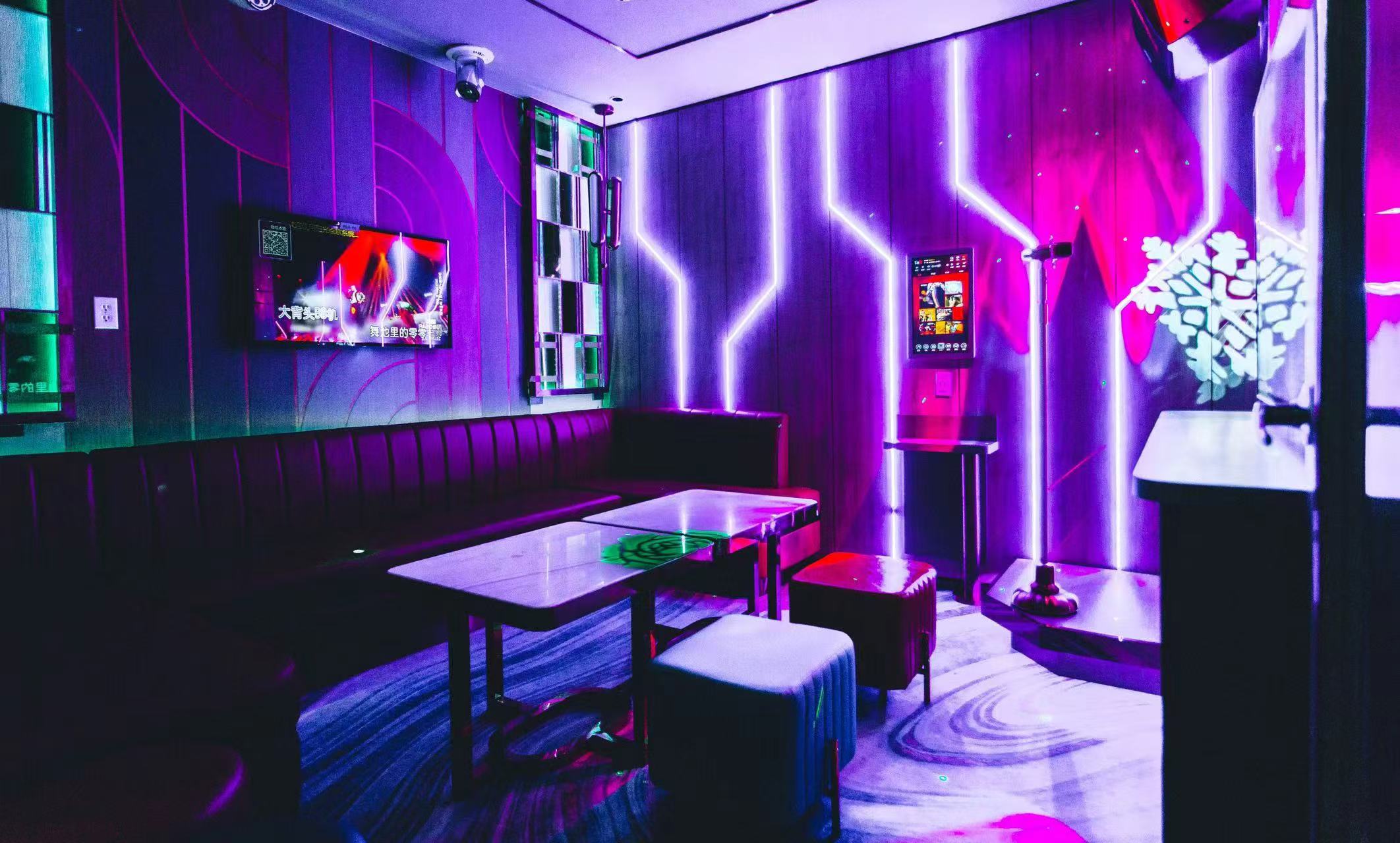 Medium karaoke room at V Show under purple lighting, with a long banquette, two tables and a wall-mounted screen